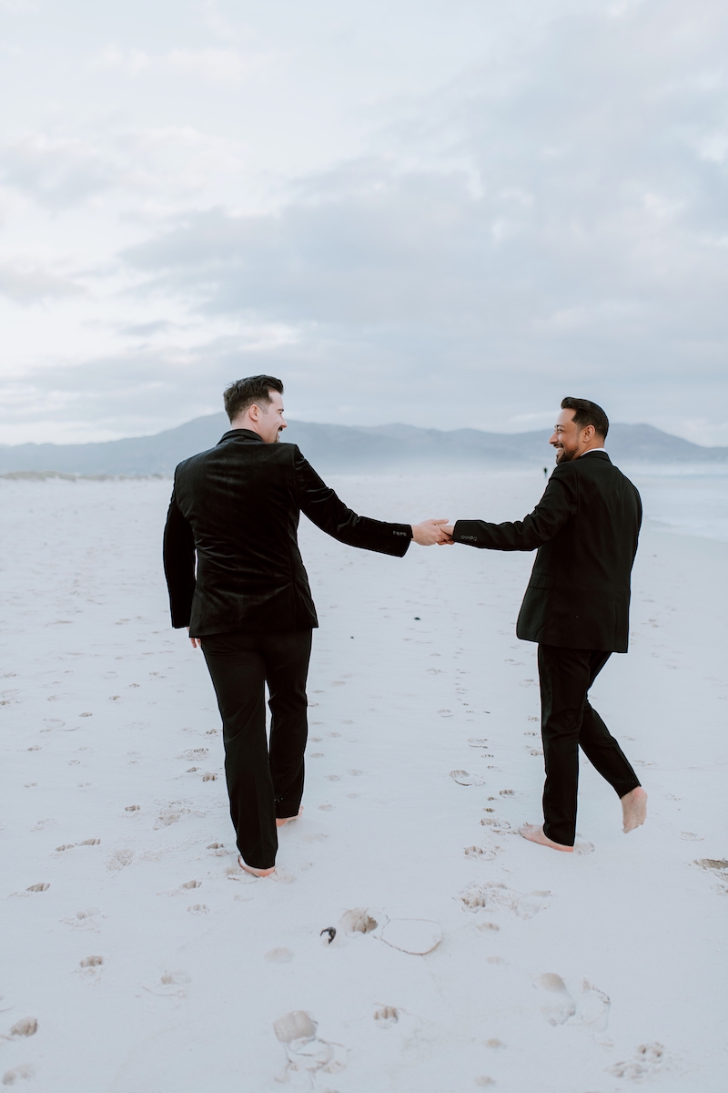 Two men in black suits walk barefoot on a sandy beach, holding hands, with mountains and cloudy sky in the background.