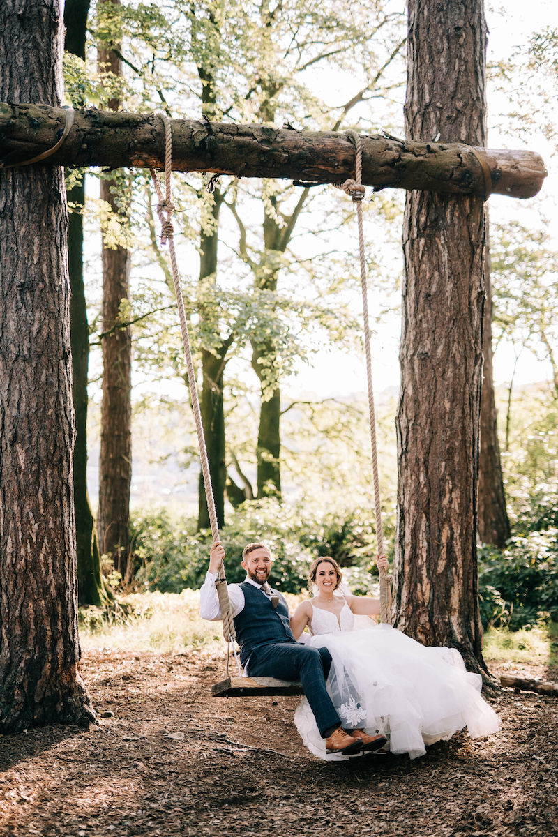 A bride and groom in wedding attire sit together on a rustic wooden swing between two trees in a forested outdoor setting.