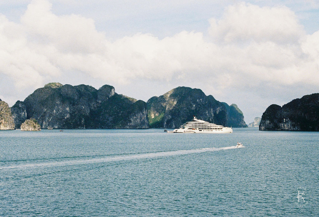 A large white cruise ship sails through calm blue water surrounded by rocky, green islands under a partly cloudy sky.