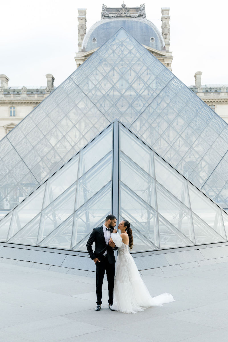 A bride and groom stand together in front of the glass pyramid at the Louvre Museum in Paris.