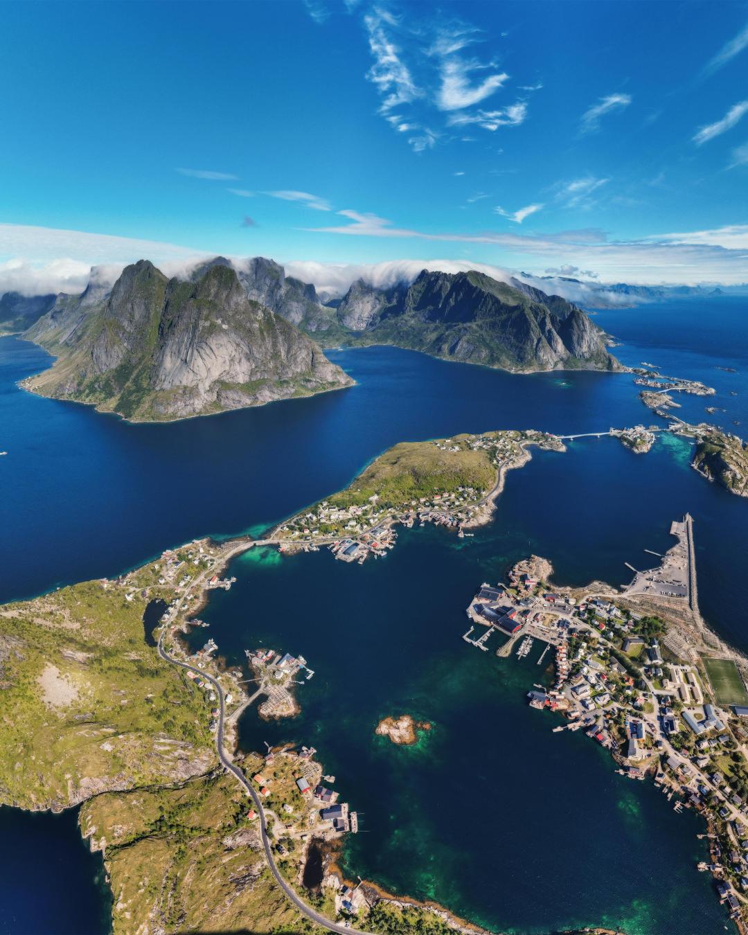 Aerial view of a coastal village surrounded by blue water, green hills, and mountains under a partly cloudy sky.
