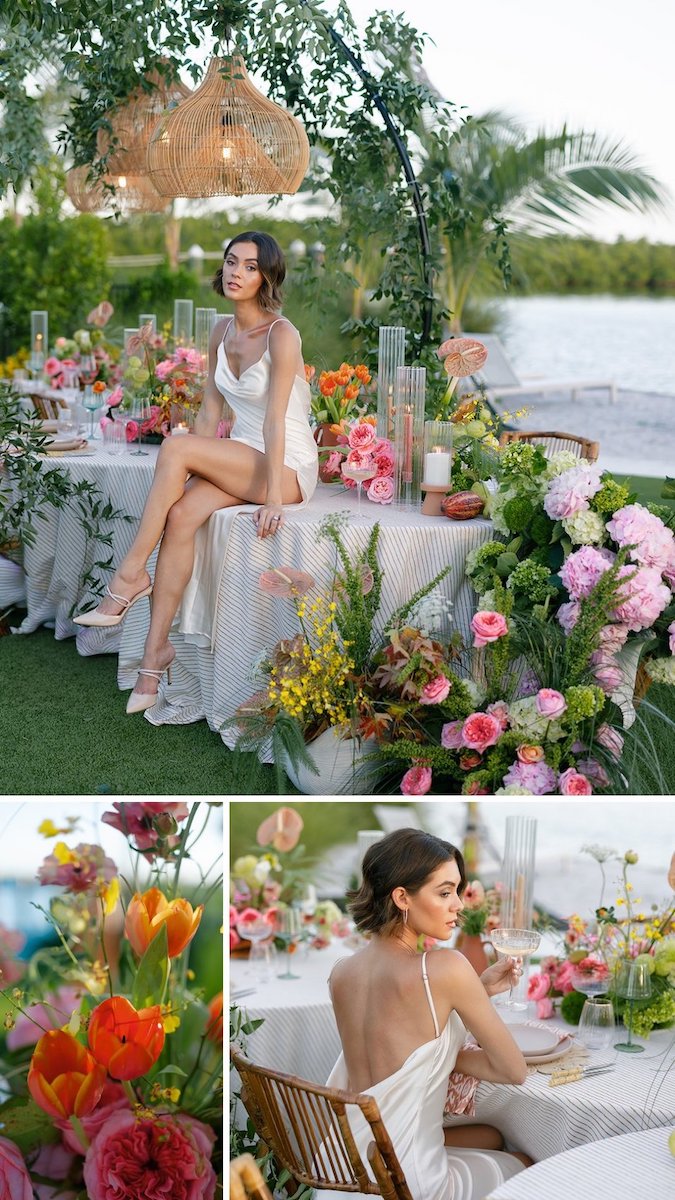 A woman in a white dress sits at a decorated outdoor table with colorful flowers and greenery by the water under hanging wicker lights.