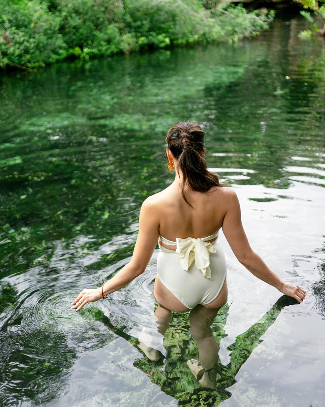 A woman in a white swimsuit stands in clear, shallow water surrounded by lush green vegetation.