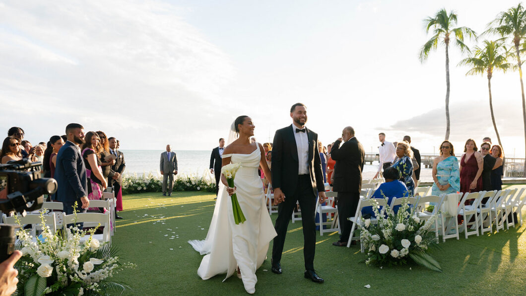 A bride and groom walk hand-in-hand down an outdoor aisle, smiling, with seated guests on either side and the ocean visible in the background.