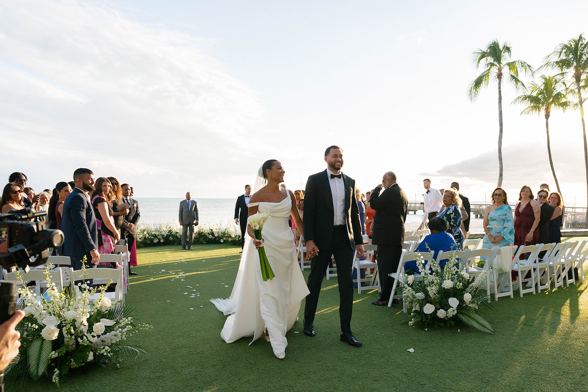 A bride and groom walk hand-in-hand down an outdoor aisle, smiling, with seated guests on either side and the ocean visible in the background.