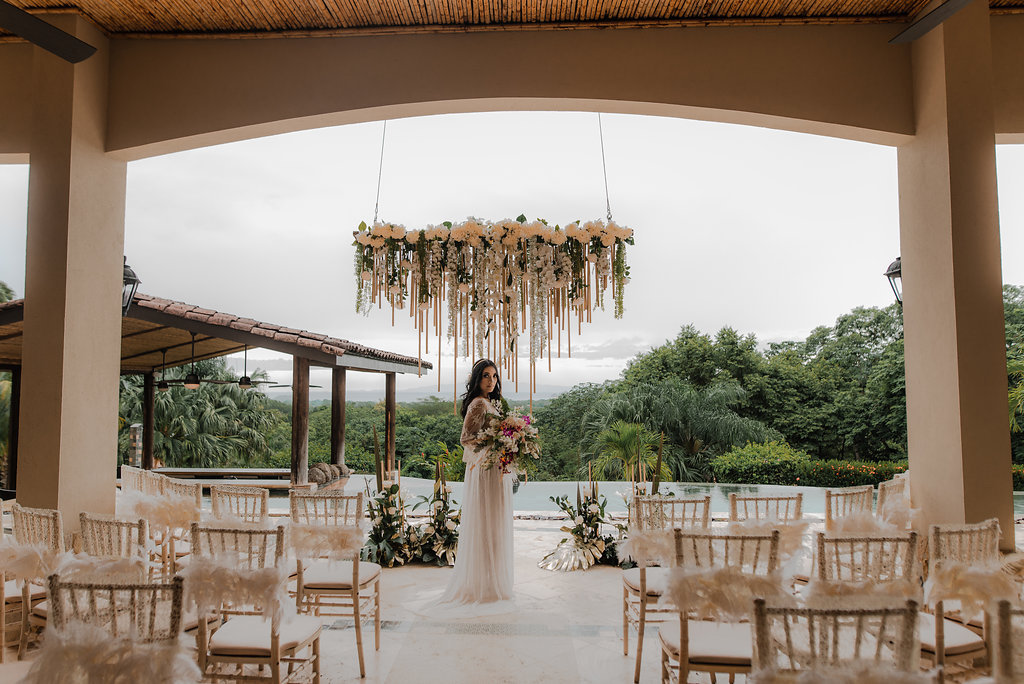 A bride in a white dress stands holding a bouquet under a floral chandelier in an outdoor wedding venue with empty decorated chairs and greenery in the background.