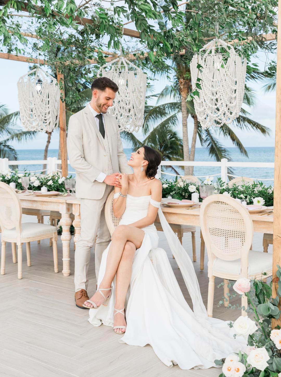 A bride in a white gown sits while a groom in a light suit stands beside her, holding her hand. They are at a decorated outdoor venue with greenery and an ocean view.