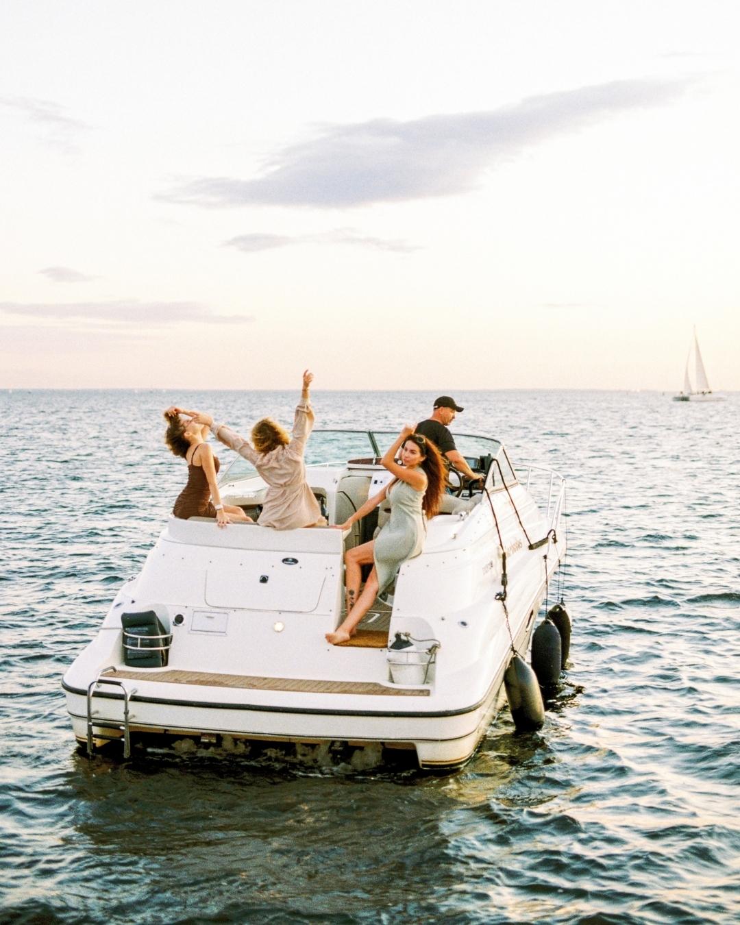Four people relax and pose on a small motorboat in open water at sunset, with one person driving and others enjoying the view.