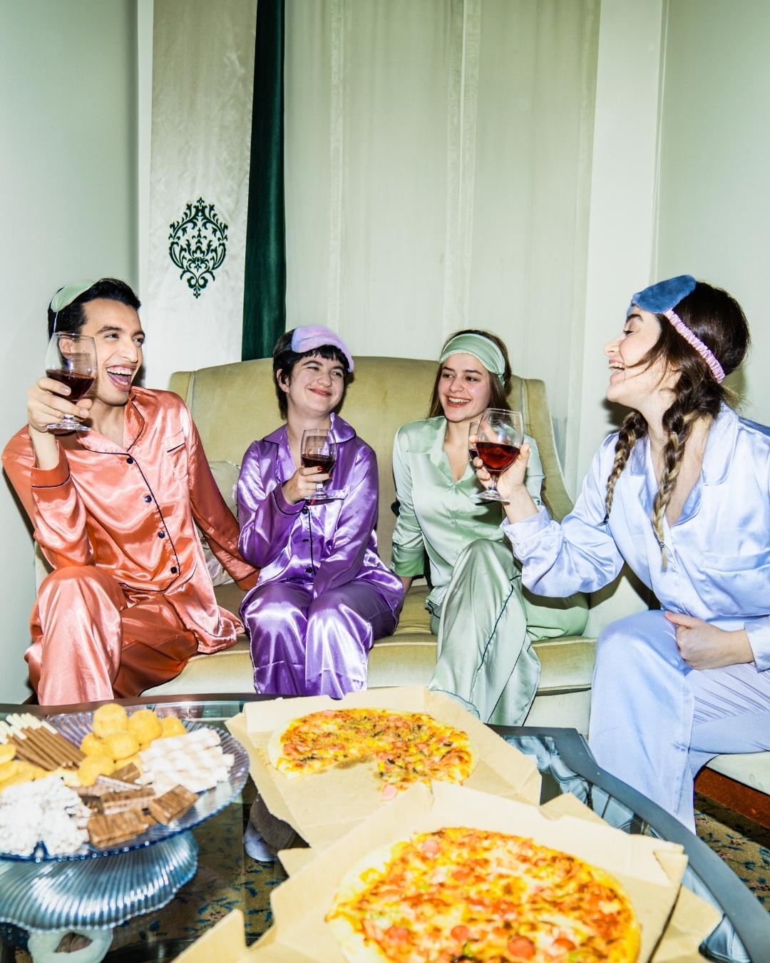 Four people in colorful pajamas sit on a couch, holding glasses of wine and smiling, with pizza and snacks on the table in front of them.