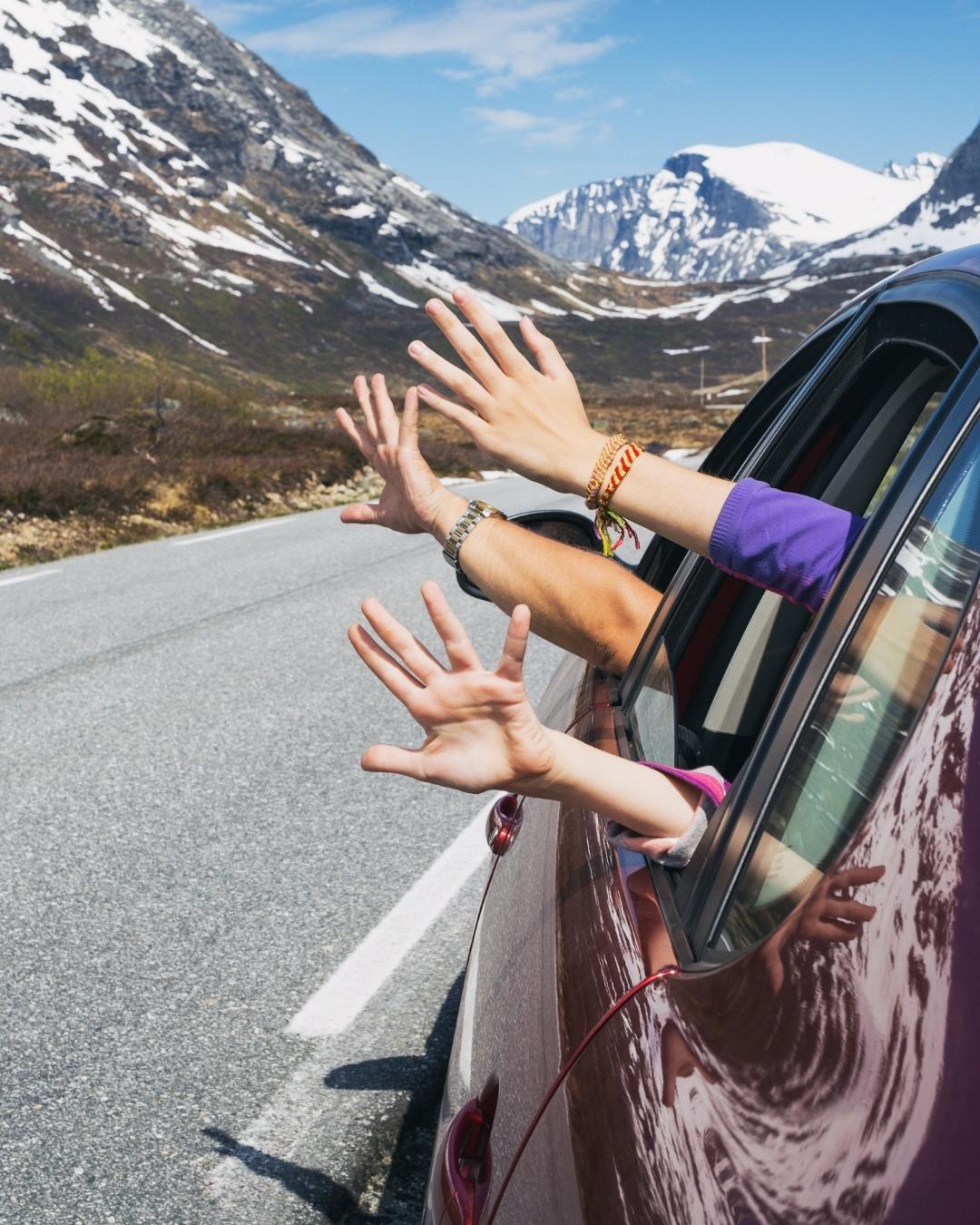 Three people wave their hands out the window of a red car driving on a mountain road with snowy peaks in the background.