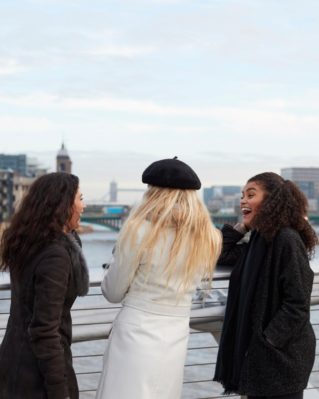 Three women stand outdoors near a railing, facing away from the camera, talking and laughing with a river and cityscape in the background.