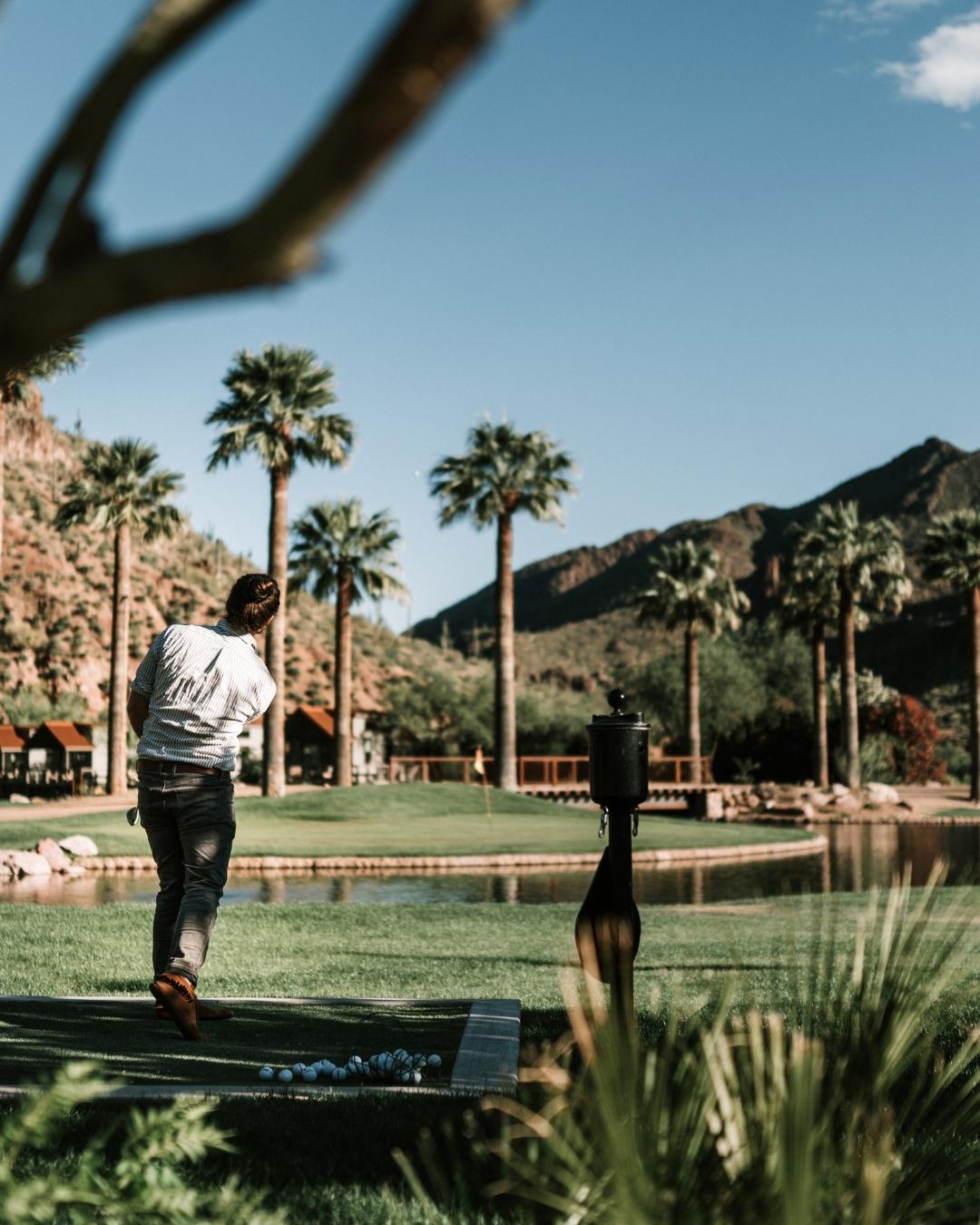 A person swings a golf club on a lush green course with palm trees, mountains, and a pond in the background under a clear blue sky.