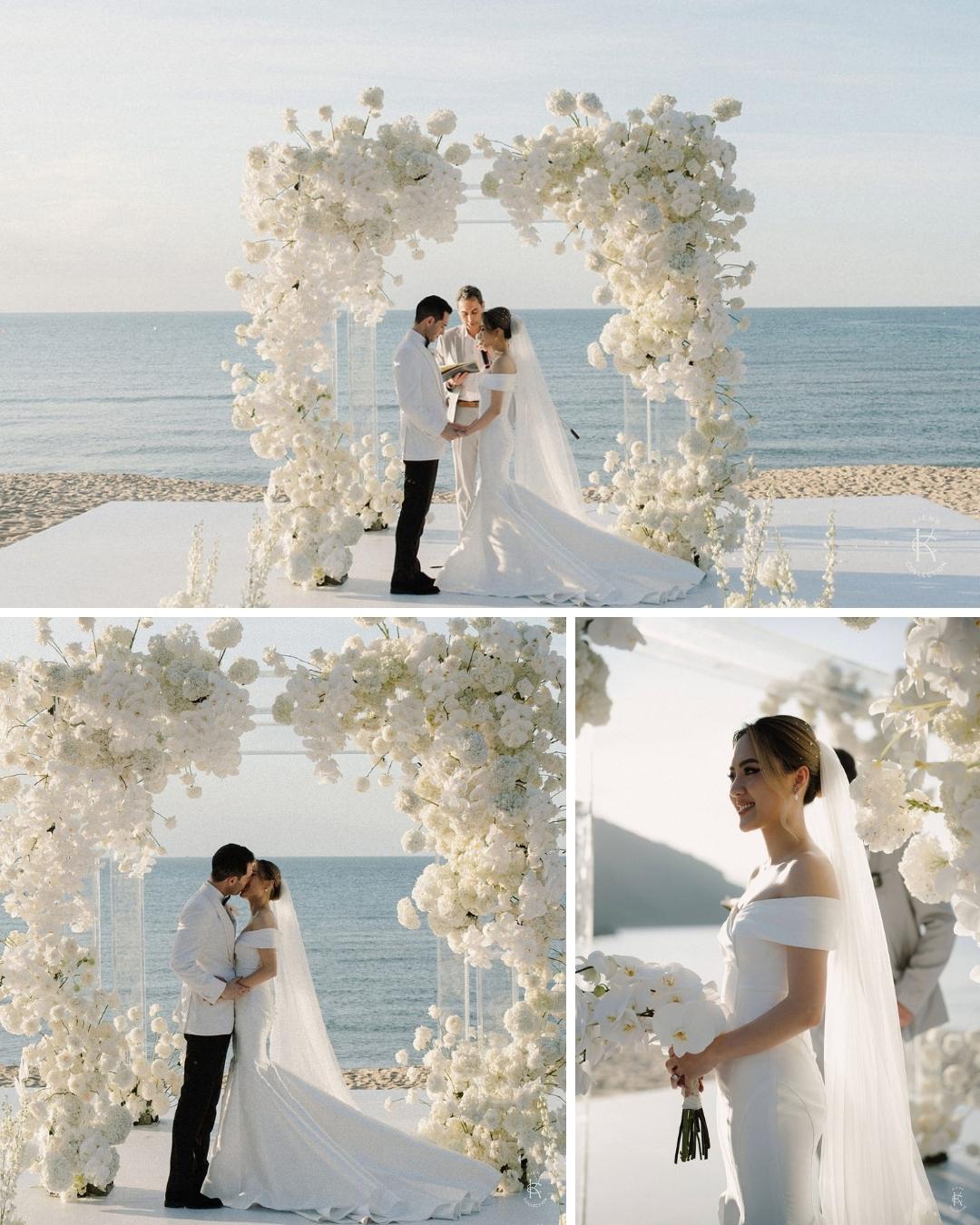 A bride and groom stand under a white floral arch by the ocean, embracing and posing for wedding photos. The bride wears a white dress and veil, holding a bouquet.