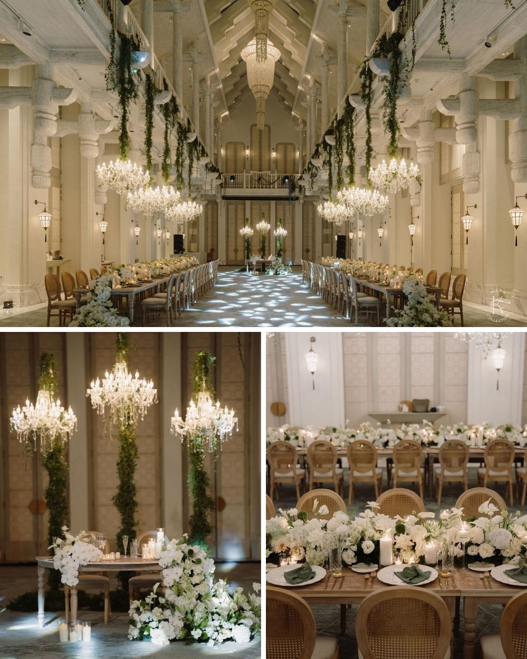 Elegant indoor wedding venue with high ceilings, chandeliers, long dining tables, white floral arrangements, and greenery accents.