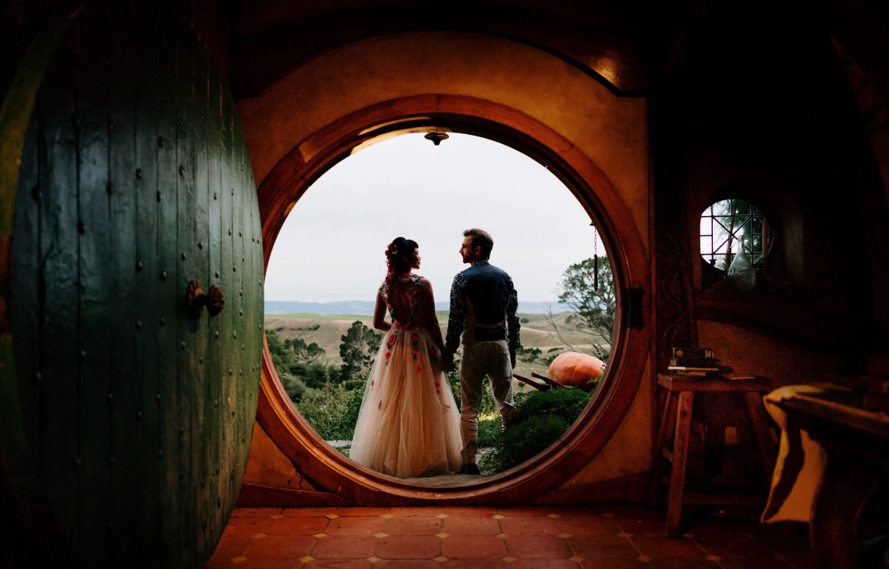 A couple stands in the doorway of a round, hobbit-style house, looking out over a scenic landscape.