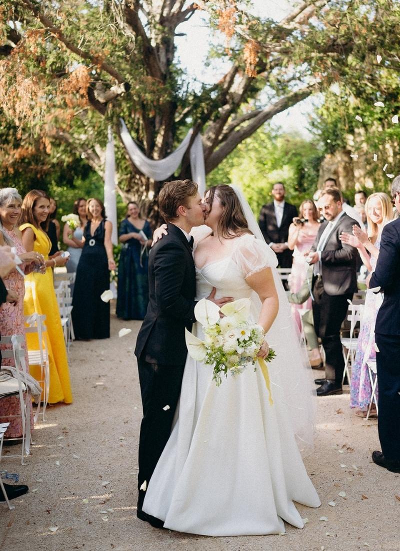 A bride and groom kiss at an outdoor wedding ceremony while guests stand and applaud around them.