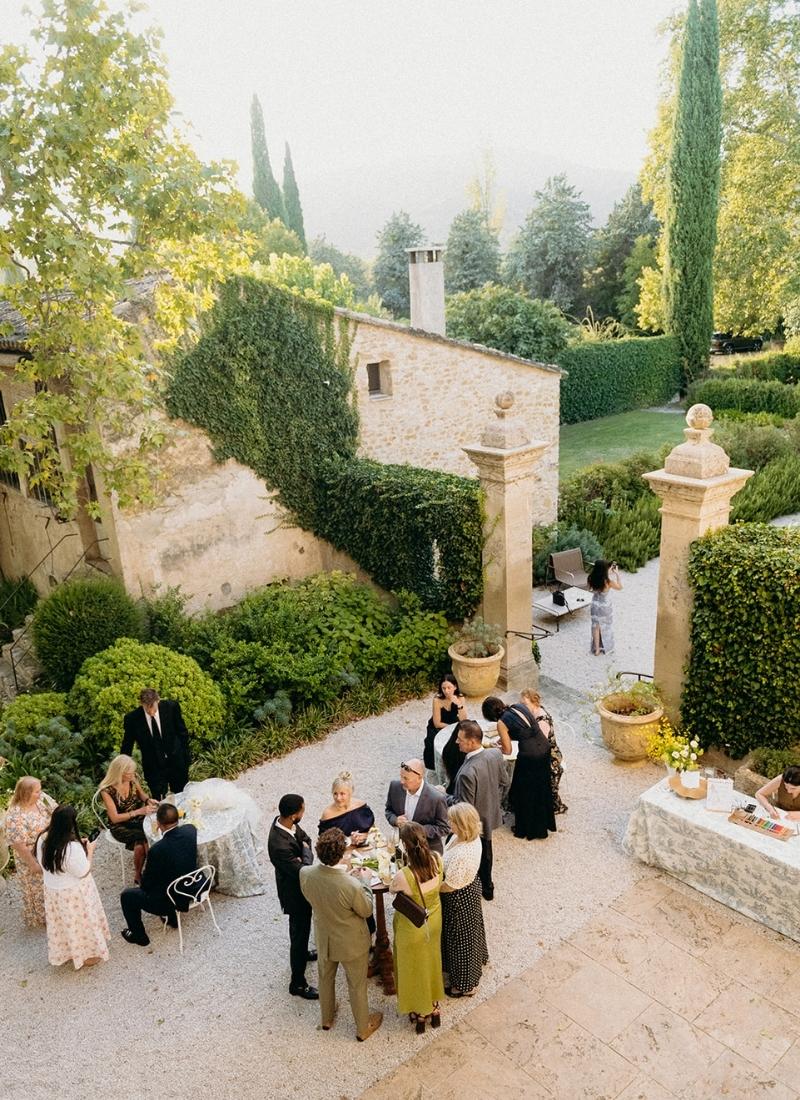 A group of people in formal attire gather around tables in an outdoor courtyard surrounded by greenery and stone buildings.