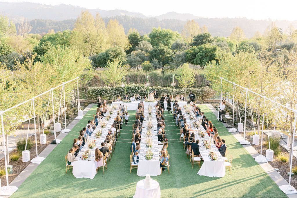 Outdoor wedding reception with long banquet tables, guests seated, string lights overhead, and lush greenery in the background.
