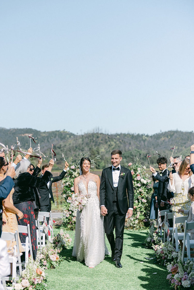 A bride and groom walk down an outdoor aisle lined with guests and flowers, with hills in the background.