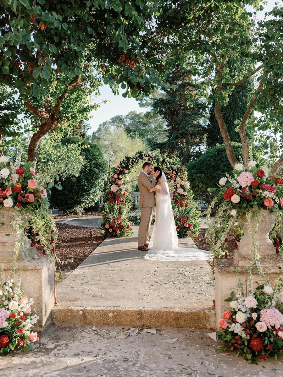 A bride and groom stand embracing on a stone pathway under a floral arch, surrounded by lush greenery and flower arrangements.