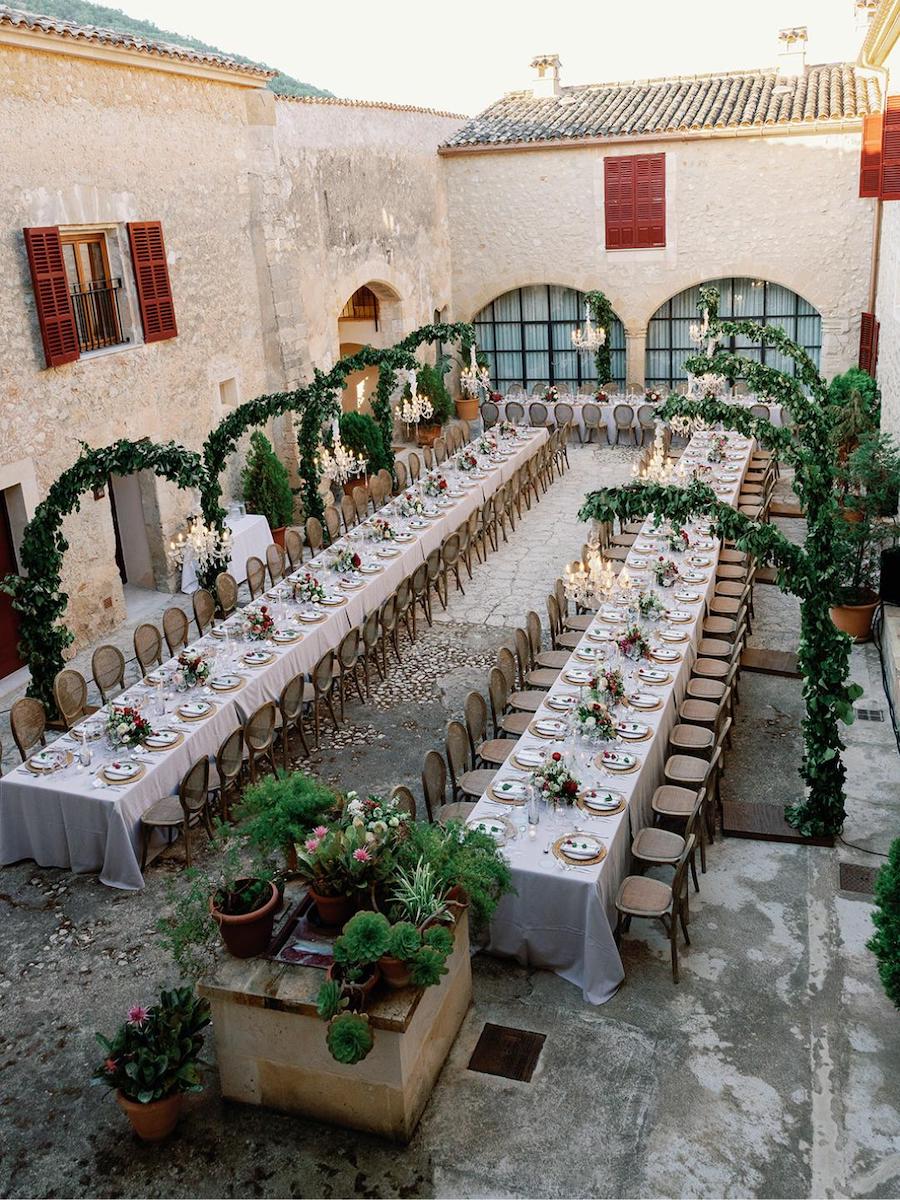 Long banquet tables set for a formal event in a rustic outdoor courtyard with stone walls, greenery arches, and potted plants.