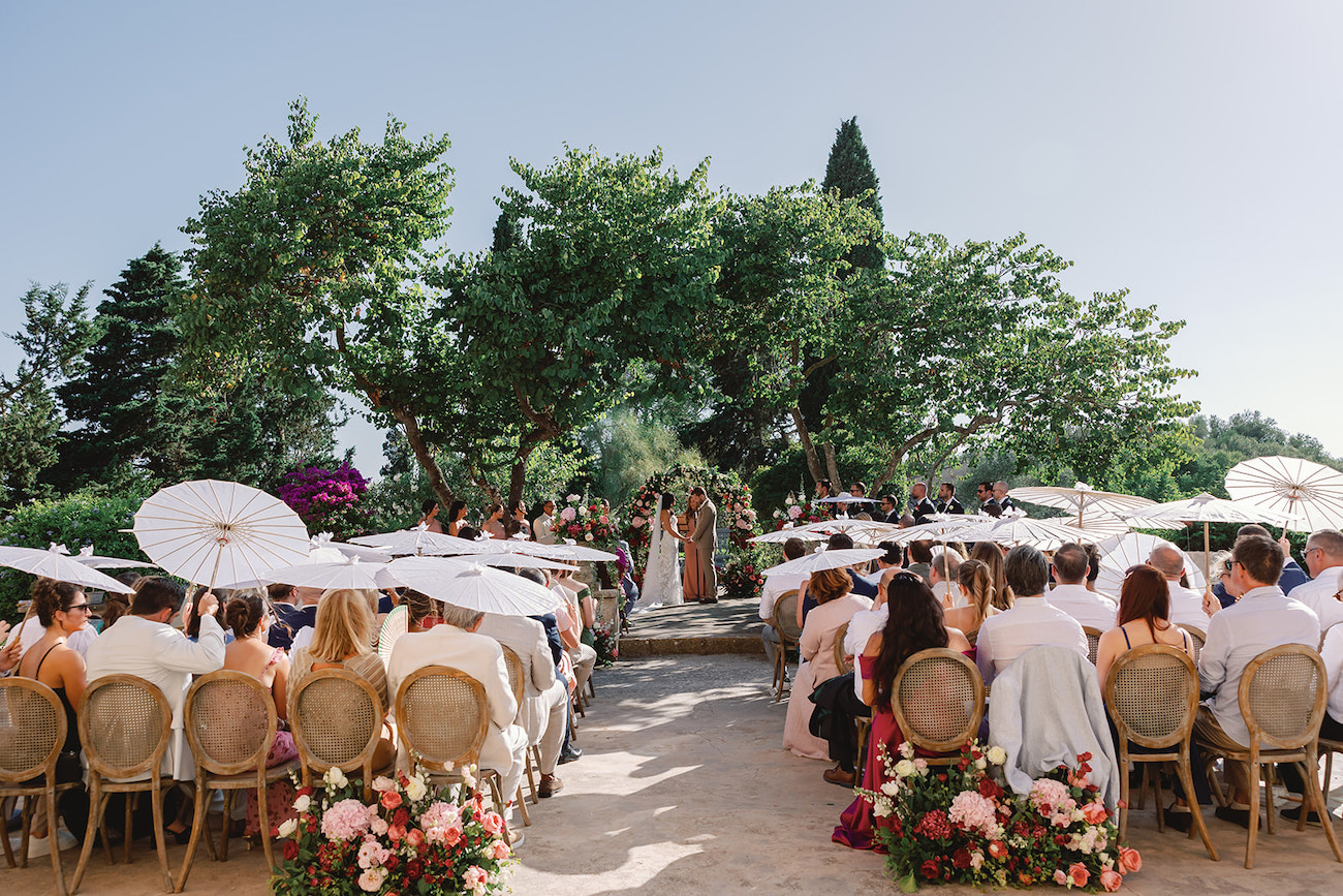 Outdoor wedding ceremony with guests seated on chairs, holding white parasols, facing a couple at the altar under trees on a sunny day.