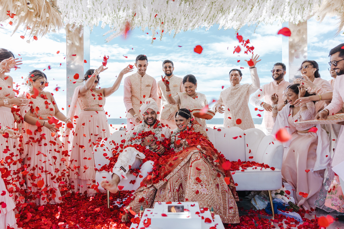 A bride and groom sit surrounded by friends as red rose petals are thrown in the air during a traditional wedding ceremony under a white floral canopy.