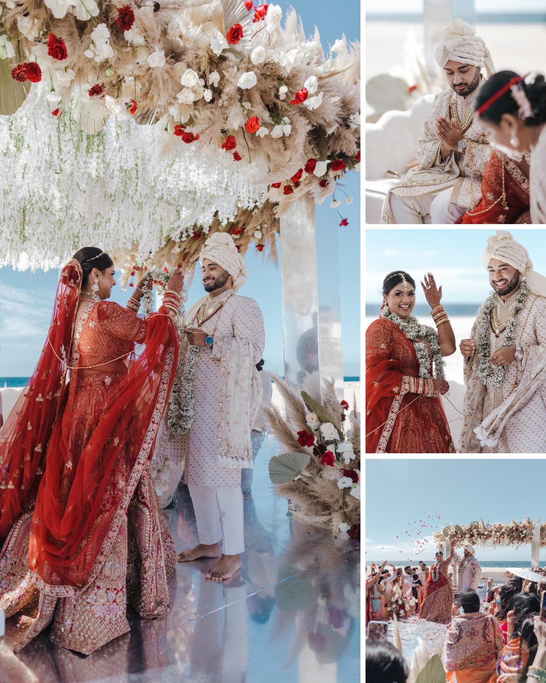 A couple in traditional Indian wedding attire takes part in a beachside ceremony under a floral canopy, smiling and exchanging garlands.