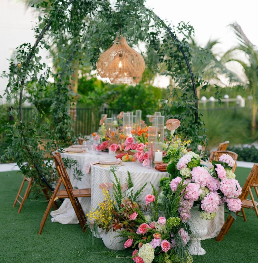 A round outdoor table with a white tablecloth is decorated with pink flowers and greenery, surrounded by wooden chairs and set under a leafy arch with a hanging woven light fixture.