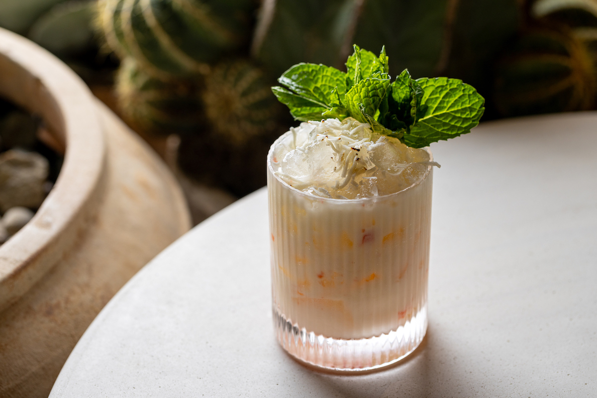 A creamy cocktail in a glass, topped with crushed ice and a fresh mint sprig, sits on a round white table with cacti in the background.