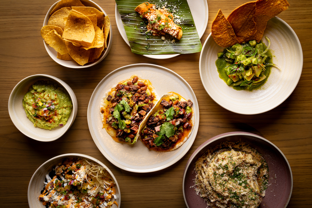 A variety of Mexican dishes on a wooden table, including tacos, guacamole, tortilla chips, a tamale, salad, and other sides served in ceramic bowls and plates.