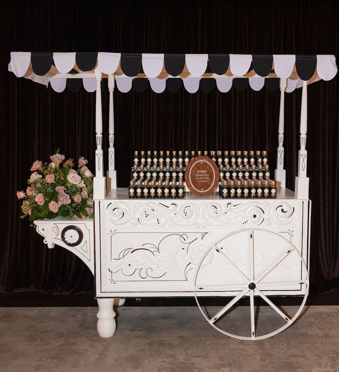 A white decorative cart with black and white striped canopy displays rows of small bottles and a round sign. Pink flowers are arranged on the left side of the cart.