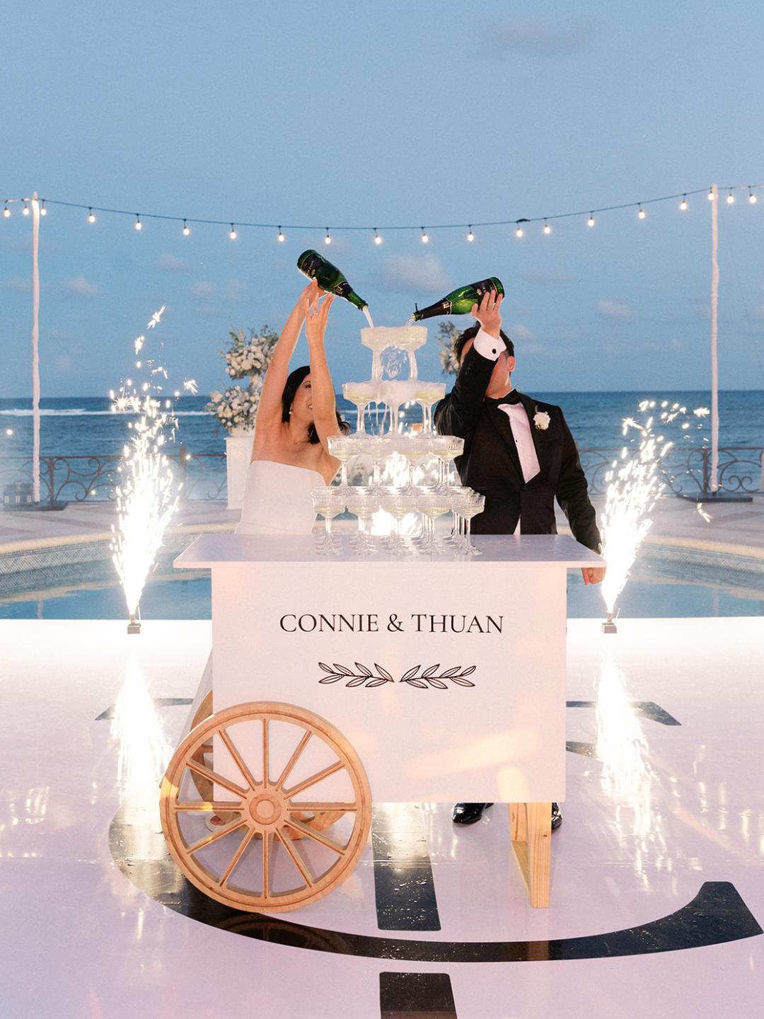 A bride and groom pour champagne into a tower at an outdoor wedding by the ocean, with sparklers and a sign reading "Connie &amp; Thuan" on the cart.