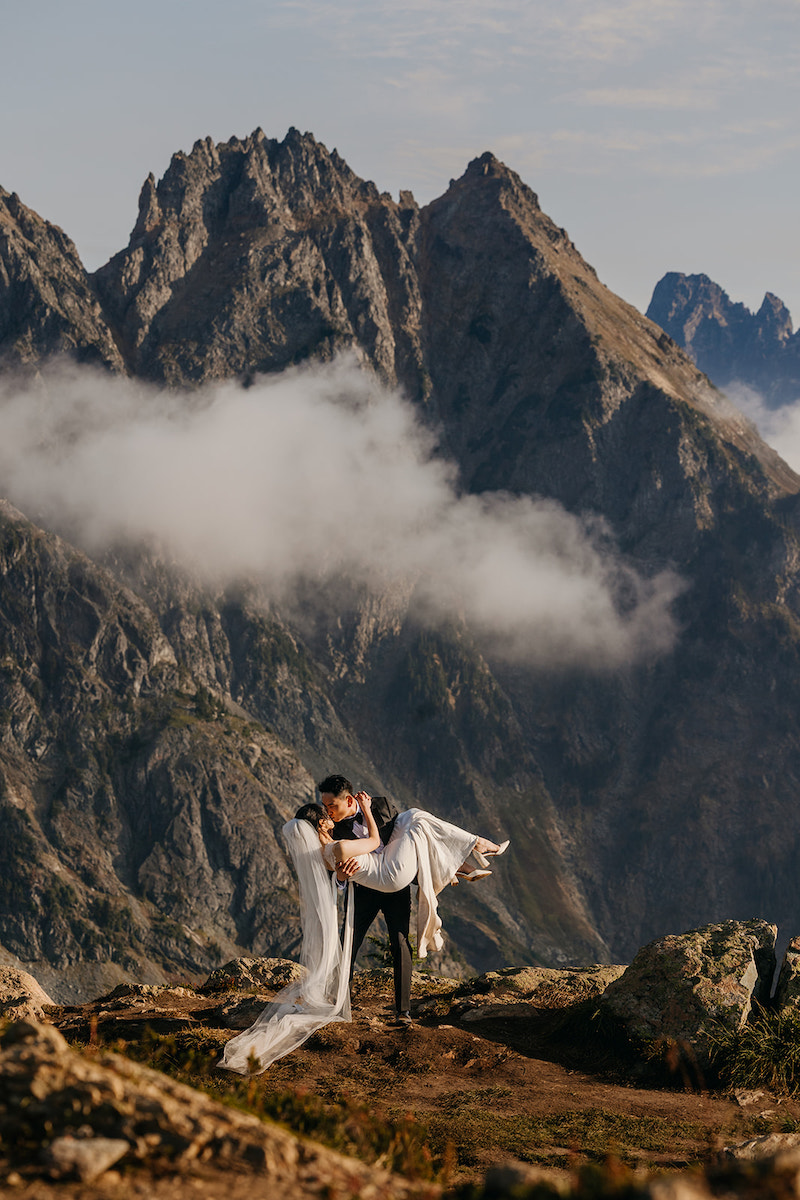 A bride and groom kiss on a rocky mountain ledge with dramatic peaks and clouds in the background.