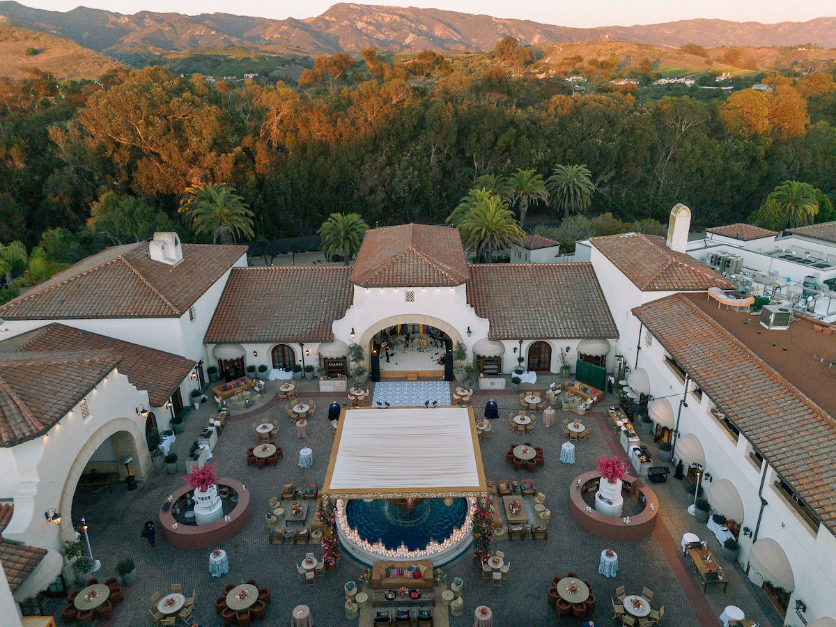 Aerial view of a large Spanish-style villa courtyard set up for an outdoor event with tables, seating, and a central canopy, surrounded by trees and hills at sunset.