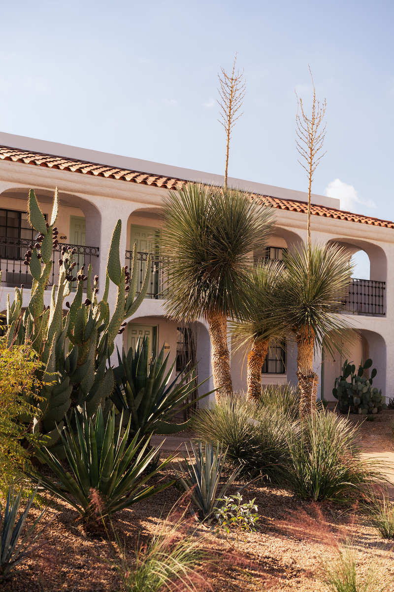 Two-story white stucco building with arches and balconies, surrounded by desert plants and cacti in the foreground.