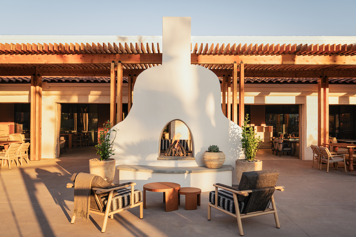 Outdoor seating area with four chairs and small tables arranged around a white stucco fireplace, set in front of a modern building with wooden accents.