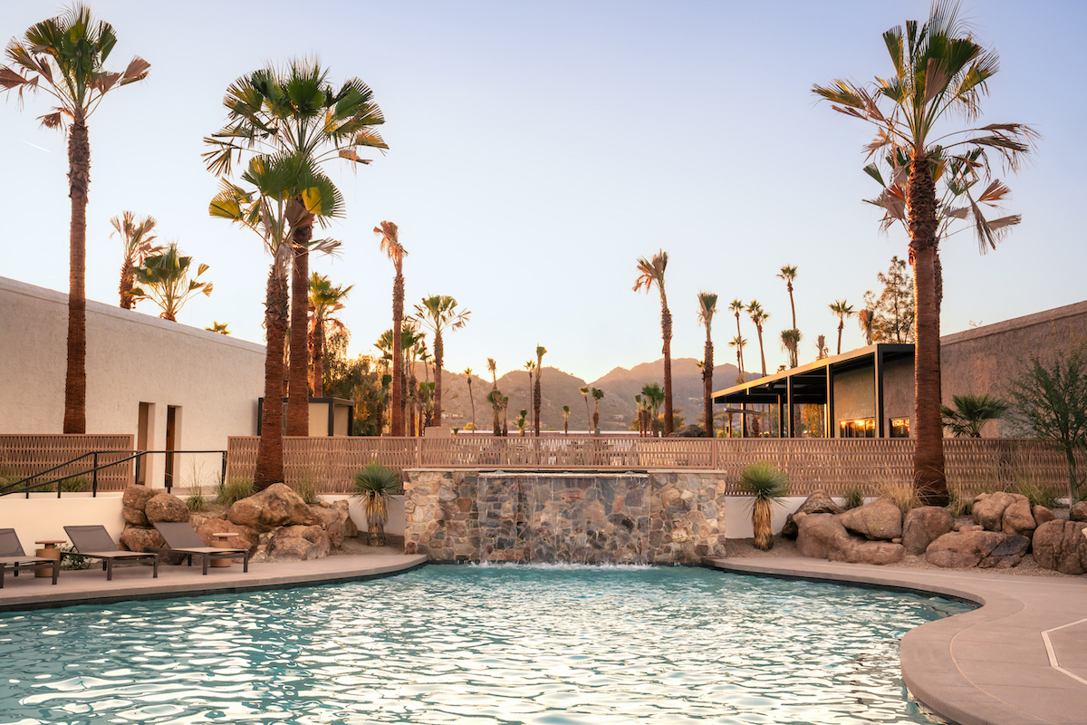 Swimming pool surrounded by lounge chairs, palm trees, and rock landscaping, with modern buildings and mountains visible in the background at sunset.