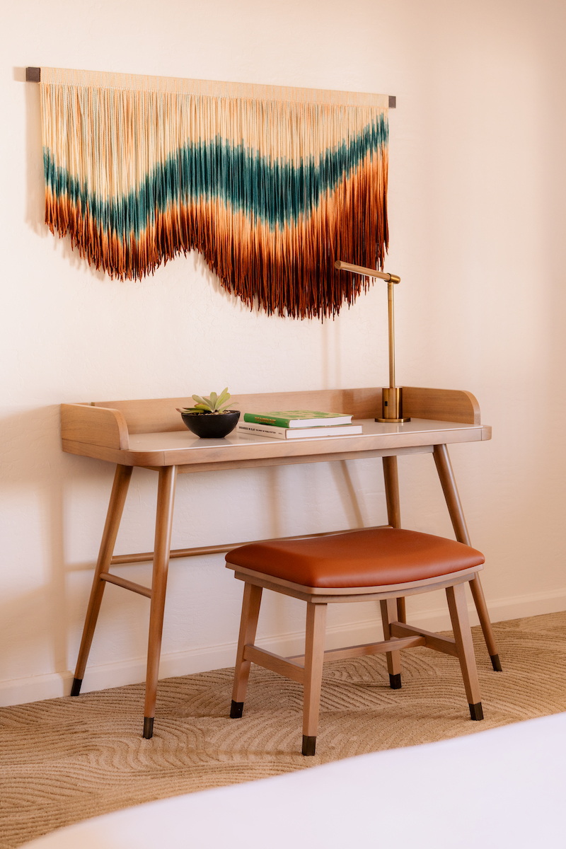 A wooden desk with a matching stool, a desk lamp, a small plant, and books, with a colorful wall hanging above.