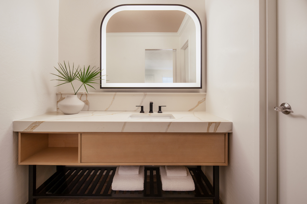A modern bathroom vanity with a large mirror, a white counter, black faucet, a bowl with a green plant, and neatly folded towels on the shelf below.