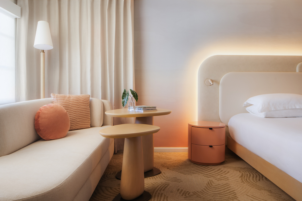 Modern hotel room with a beige sofa, round cushions, small wooden tables, a bed with a lighted headboard, and a nightstand. Neutral color palette with peach accents.