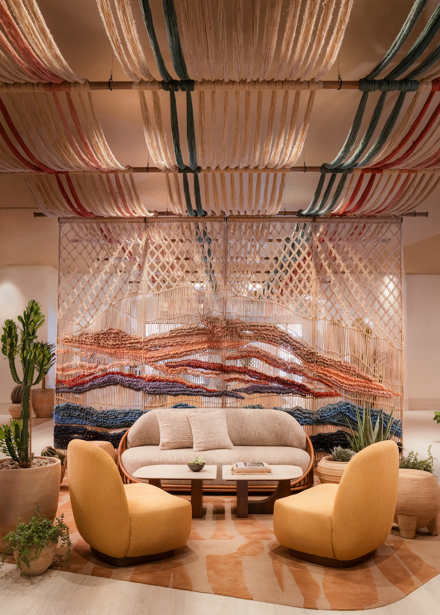A modern lounge area with a beige sofa, two yellow chairs, a round table, potted plants, and woven textile art on the wall and ceiling.