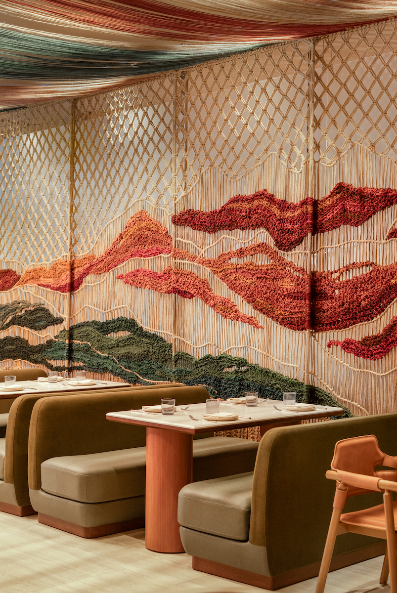 A restaurant interior with green booths, light wooden tables, a wooden chair, and a woven wall mural featuring abstract mountain designs in red, orange, and green tones.