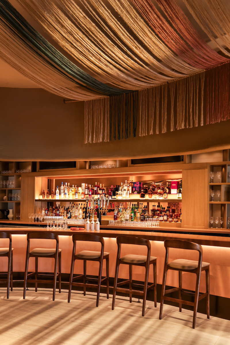A modern bar with a curved counter, padded bar stools, bottles of alcohol on display, and decorative fabric draped across the ceiling.