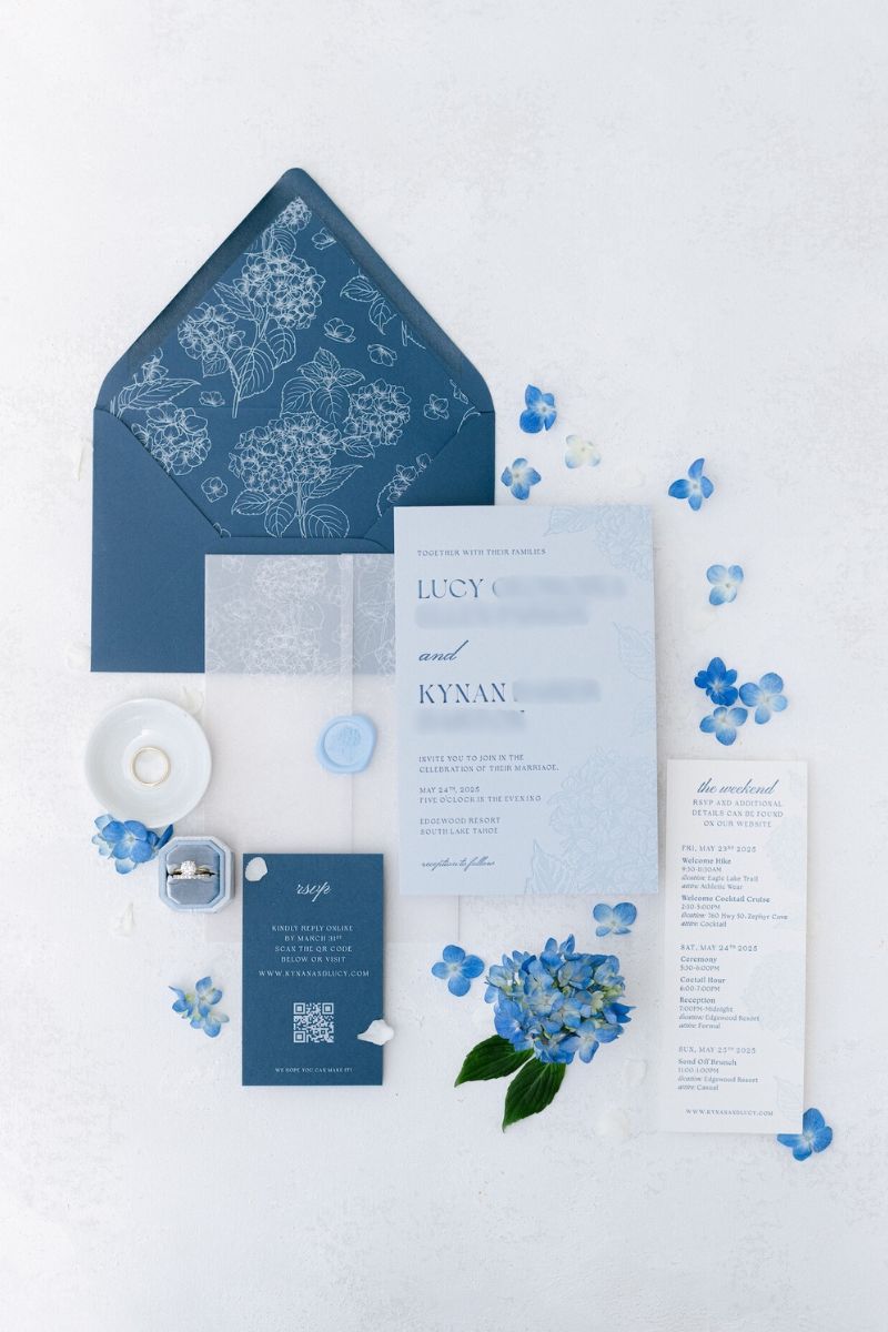 Flat lay of a blue and white wedding invitation suite with envelopes, cards, a menu, a candle, a ring box, and blue hydrangea flowers on a white background.