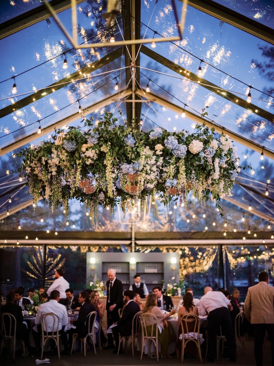 Guests sit at tables under a clear tent with string lights and a large hanging floral arrangement at an evening event.