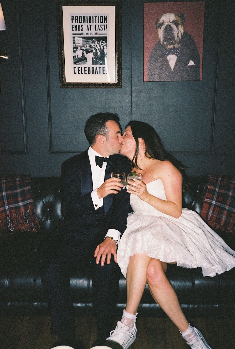 A couple dressed in formal attire sits on a black couch, kissing and holding drinks, with framed art on the wall behind them.