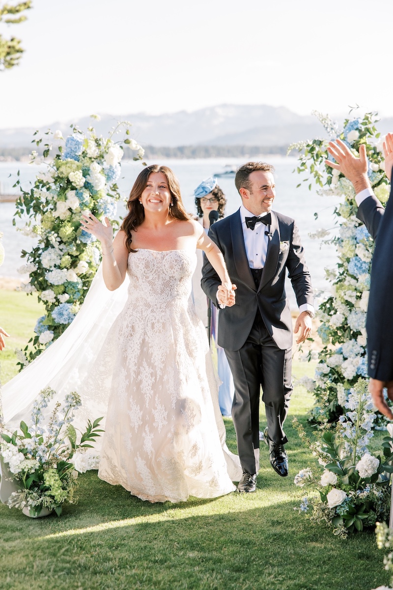 A bride and groom walk hand-in-hand outdoors, smiling and waving, surrounded by floral arrangements with a lake and mountains in the background.