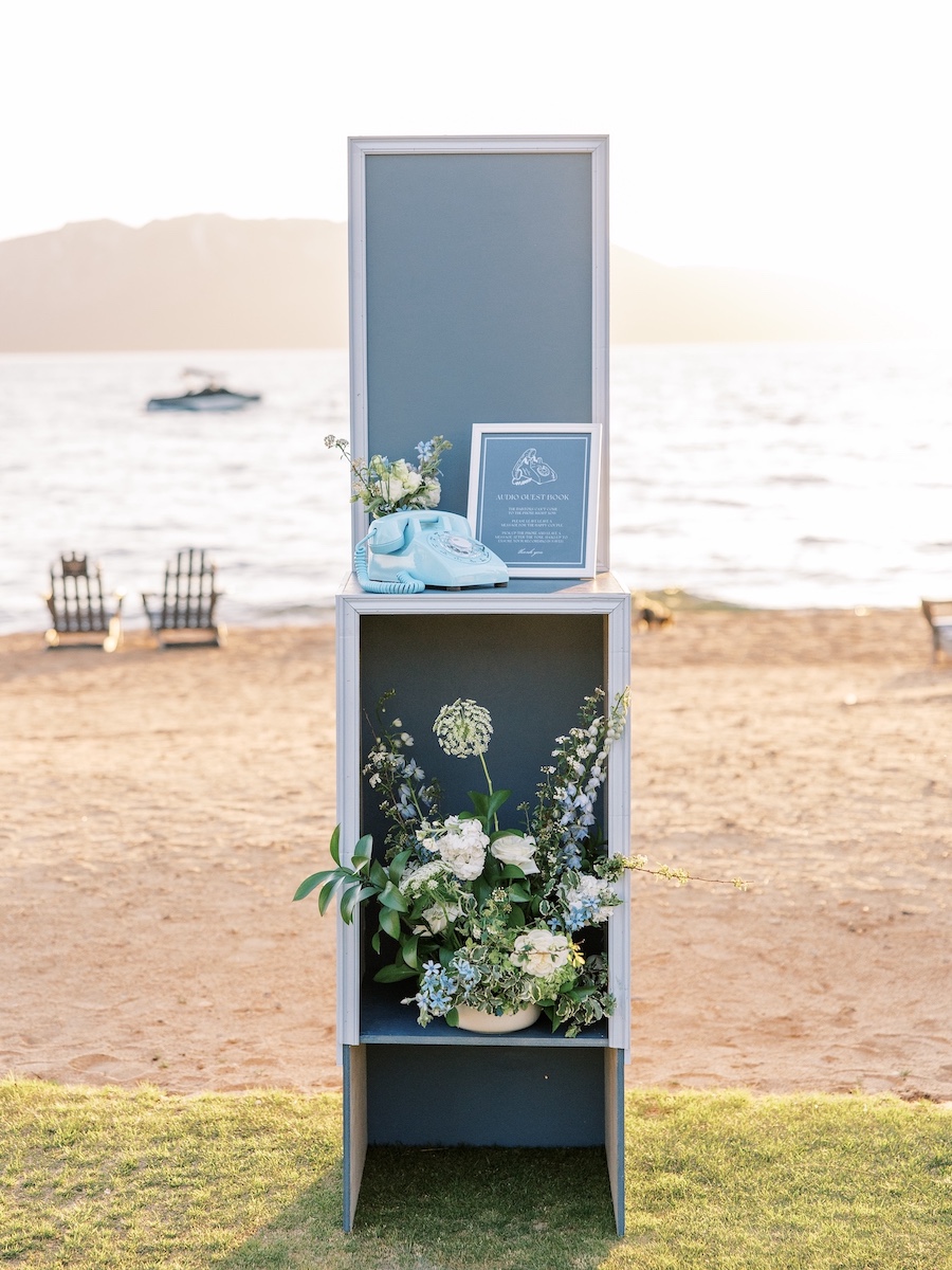 A decorative display with flowers and a framed sign stands on the sand by the water, with mountains and chairs visible in the background.