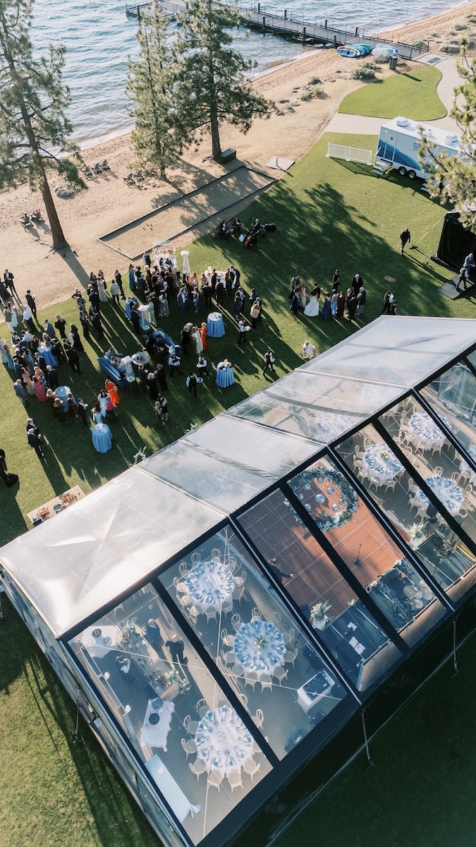 Aerial view of an outdoor event by a lake, with people gathered on grass near a tented area set for dining and trees providing shade.
